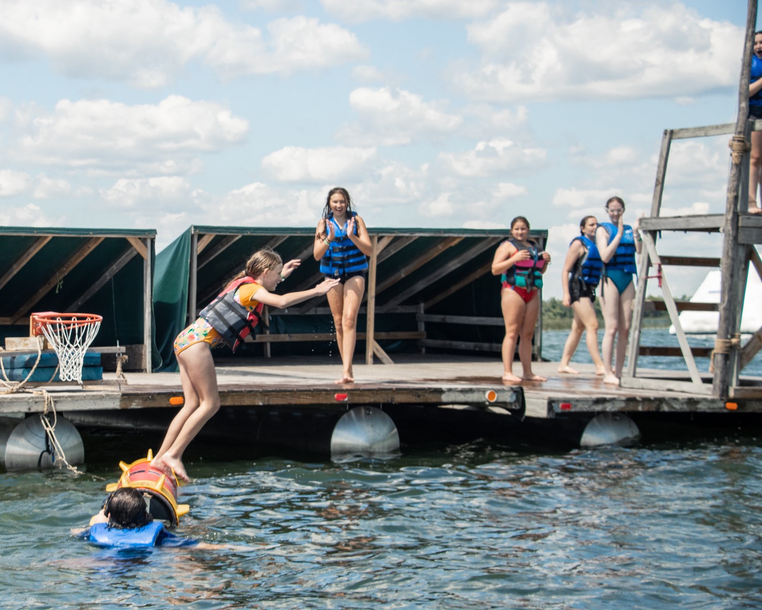 A group of Scouts are wearing life jackets and playing on a large raft in the lake. The raft is made with large wooden beams and is floating by using pontoon rafts.