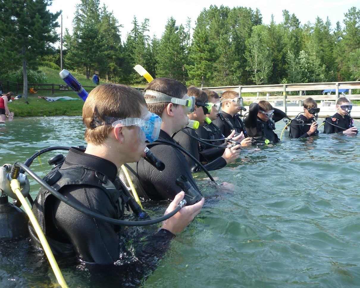 A group of Scouts wearing full SCUBA gear (goggles, air tank, breather, etc.) are watching the instructor