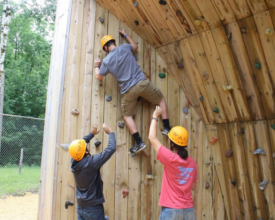 An older Scout bouldering up the bouldering wall as two other older Scouts act as spotters