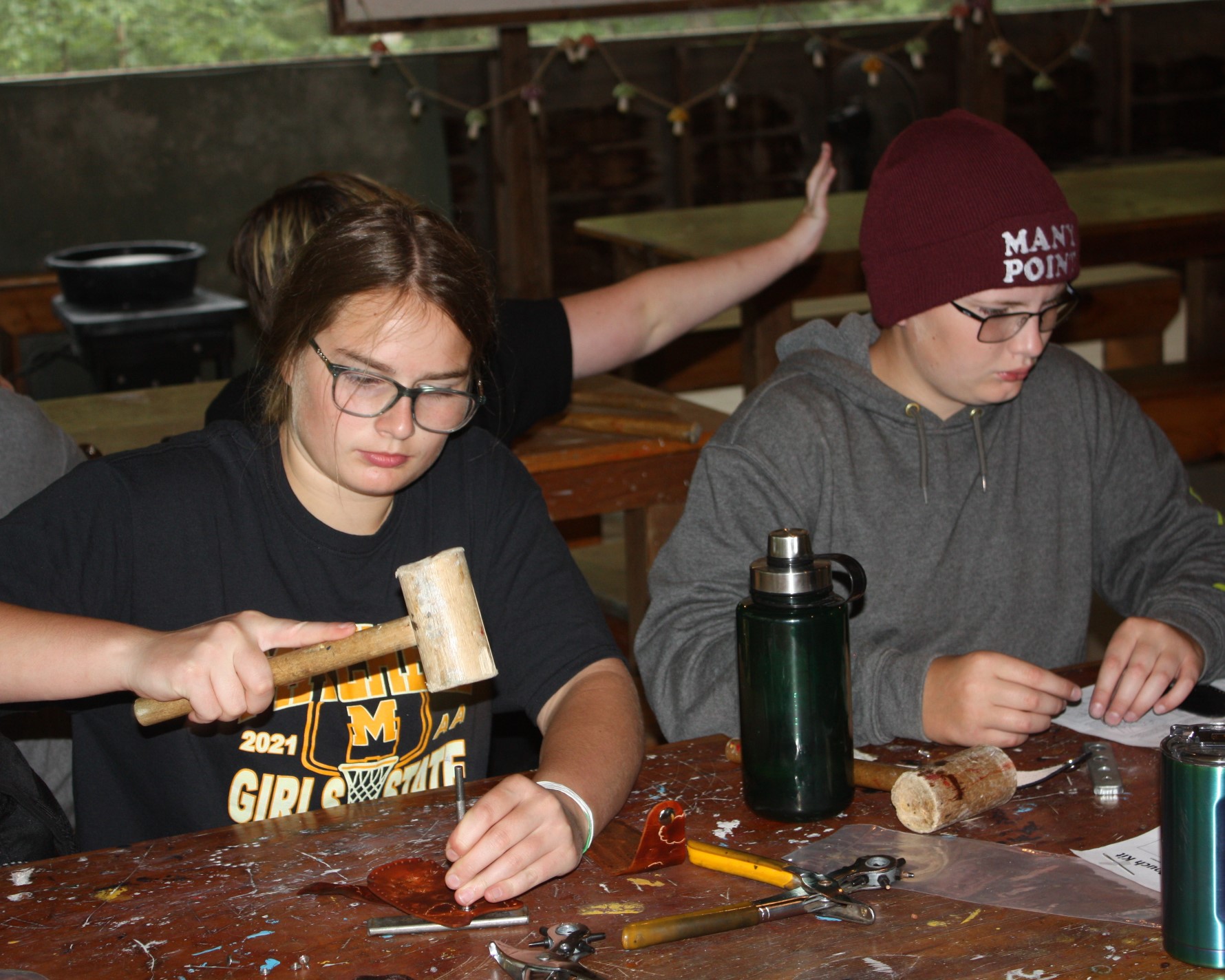 Two Scouts working on leather projects. One is hammering a metal stamp into a small leather cutout on the table, the other is tracing a design into a piece of paper