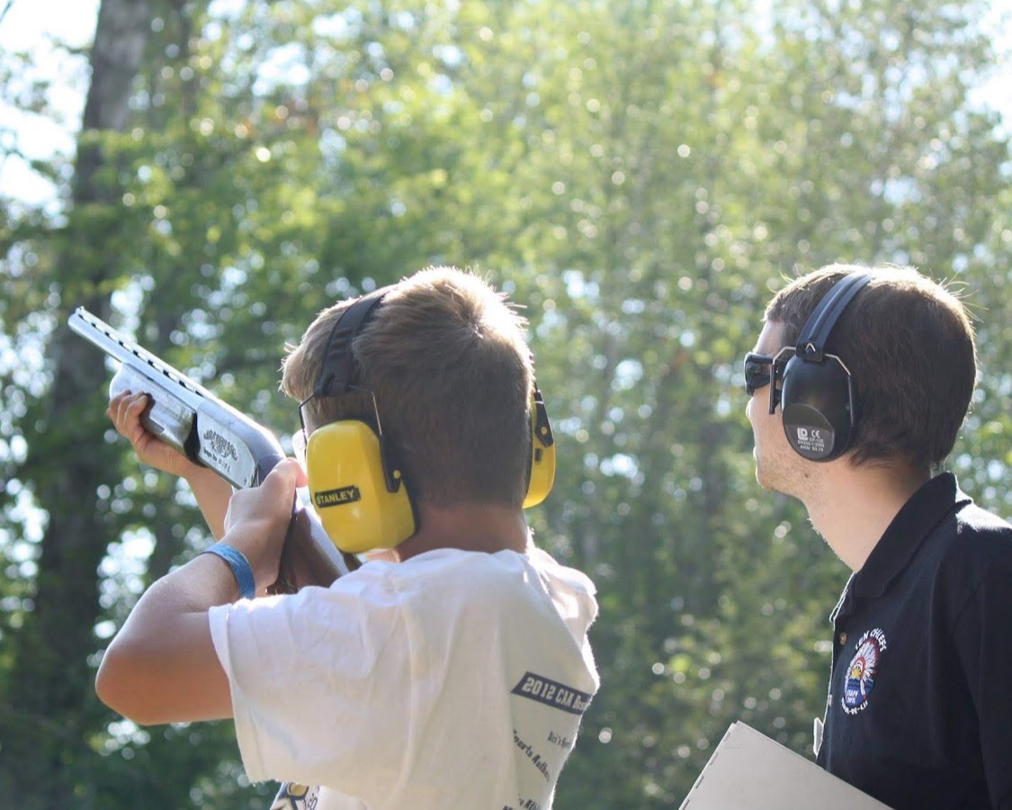 A Scout is aiming the shotgun waiting for the clay pigeon to be fired while a staff member watches