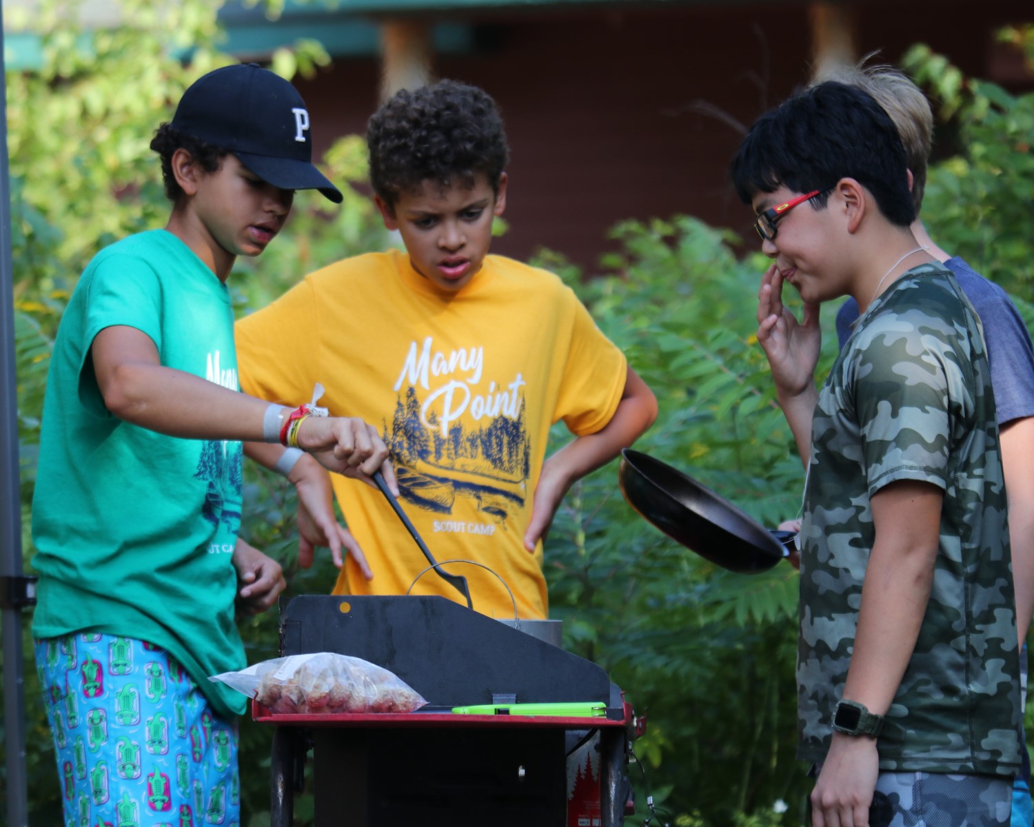 Four Scouts wearing Many Point t-shirts surround a table with a camp stove on it, all doing some preparation work for cooking.