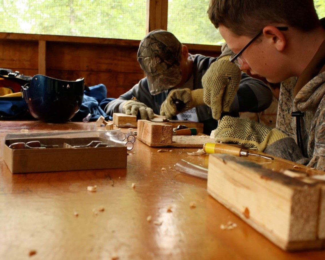 Two Scouts looking down and carving a block of wood into some unknown project